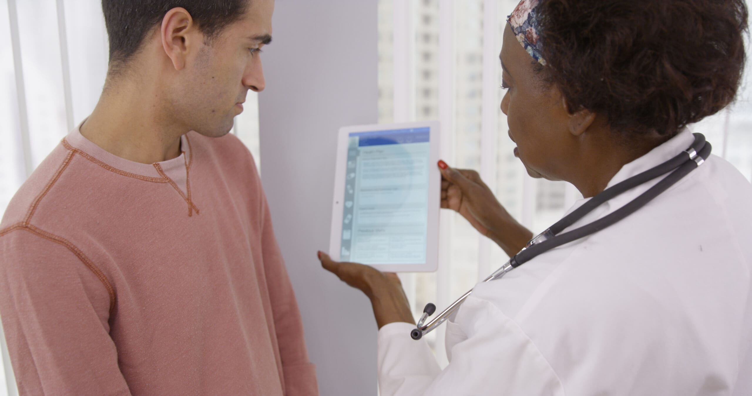 A healthcare provider in a white coat shows a patient a digital tablet with health information, engaging him in a discussion. The provider's stethoscope rests on her neck, emphasizing a focus on care and communication.