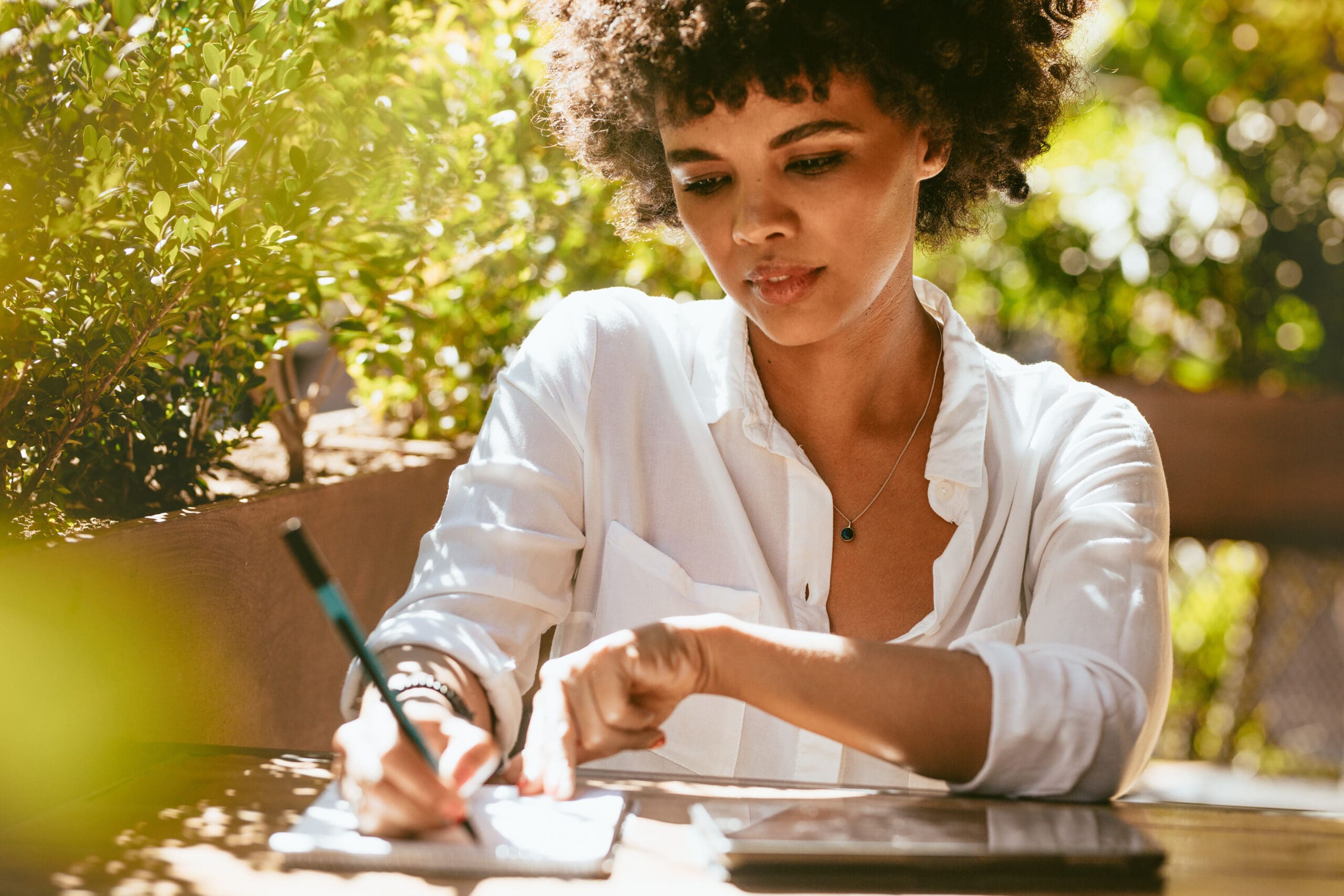 Freelancer doing her work at outdoors coffee shop. African female writing on notepad sitting at a coffee table.